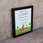 Wall art with a green field, trees, and cartoon figures of Jesus and children, featuring the text 'Believe in the Lord Jesus Christ, and you will be saved. displayed on the wall in a black frame