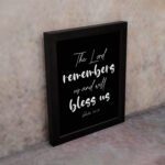 The Lord Remembers Us And Will Bless Us - Psalm 115:12 Bible Verse Wall Art on a black background with white text. displayed on the wall in a black frame