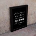 Acts 2:21 Bible wall art featuring bold white text on a black background, reading And everyone who calls on the name of the Lord will be saved. displayed on the wall in a black frame