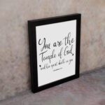 You Are The Temple Of God" Bible Wall Art with gold text on a black background from 1 Corinthians 3:16. displayed on the wall in a black frame