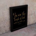 You Are The Temple Of God" Bible Wall Art with gold text on a black background from 1 Corinthians 3:16. displayed on the wall in a black frame