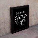 Black wall art with white text reading 'I am a CHILD of god' from John 1:12, showcasing a mix of cursive and bold lettering. displayed on the wall in a black frame