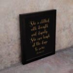 Proverbs 31:25 Christian Wall Art - She Is Clothed With Strength And Dignity, Gold Text on Black Background displayed on the wall in a black frame