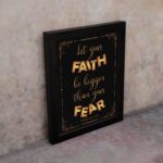 Infidu wall art with the text "Let your faith be bigger than your fear" in gold and bronze, with the Bible verse Hebrews 13:6 on a black background. displayed on the wall in a black frame