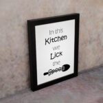 In This Kitchen We Lick the Spoon kitchen wall art featuring black text on a silver-gray background with a minimalist spoon illustration. displayed on the wall in a black frame