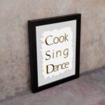 Cook Sing Dance kitchen wall art with black and orange text on a light gray background with jagged white accents. displayed on the wall in a black frame