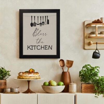 Bless This Kitchen wall art with black kitchen utensils on a beige background in cursive and bold block fonts. There is a vegetable table, and above it, there is wall art.