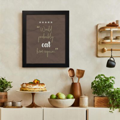 Would Probably Eat Here Again kitchen wall art with a brown background, gold and white text, and star icons for a review-inspired design. There is a vegetable table, and above it, there is wall art.