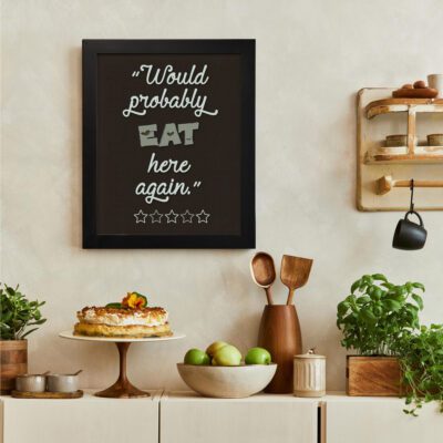 Would Probably Eat Here Again kitchen wall art with a dark brown background, bold white text in a playful layout, and five outlined stars. displayed on the wall in a black frame in a white background displayed on the table