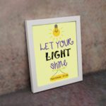 Infidu wall art featuring a lightbulb and the verse Let Your Light Shine from Matthew 5:16 in colorful fonts on a yellow background. displayed on the wall in a white frame