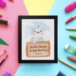 A cute white bear holding a wooden sign with the verse We Love Because He First Loved Us - 1 John 4:19 against a blue sky with clouds. displayed on the wall in a black frame in a colorful background