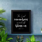 The Lord Remembers Us And Will Bless Us - Psalm 115:12 Bible Verse Wall Art on a black background with white text. displayed on the wall in a black frame