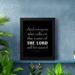 Acts 2:21 Bible wall art featuring bold white text on a black background, reading And everyone who calls on the name of the Lord will be saved. displayed on the wall in a black frame