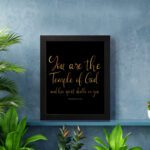 You Are The Temple Of God" Bible Wall Art with gold text on a black background from 1 Corinthians 3:16. displayed on the wall in a black frame