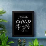 Black wall art with white text reading 'I am a CHILD of god' from John 1:12, showcasing a mix of cursive and bold lettering. displayed on the wall in a black frame