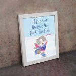 Wall art featuring a joyful bear in winter clothes holding a heart, with the verse We love because He first loved us from 1 John 4:19. displayed on the wall in a white frame