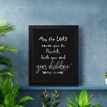 Wall art featuring the quote 'May the Lord cause you to flourish, both you and your children' from Psalm 115:14 on a black background. displayed on the wall in a black frame