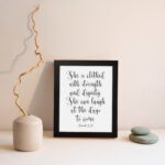 Proverbs 31:25 Christian wall art featuring the text She is clothed with strength and dignity; she can laugh at the days to come. displayed on the wall in a black frame