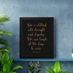 Proverbs 31:25 Christian Wall Art - She Is Clothed With Strength And Dignity, Gold Text on Black Background displayed on the wall in a black frame