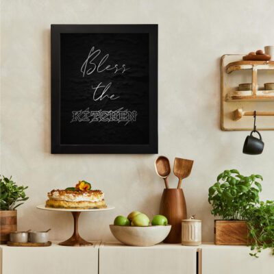 Bless This Kitchen wall art with cursive and bold fonts on a dark black textured background. There is a vegetable table, and above it, there is wall art.