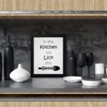 In This Kitchen We Lick the Spoon kitchen wall art featuring black text on a silver-gray background with a minimalist spoon illustration. displayed on the table