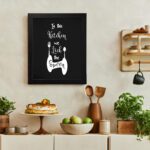 In This Kitchen We Lick The Spoon wall art with white cursive and bold fonts, black background, and stylish cutlery illustrations. There is a vegetable table, and above it, there is wall art.