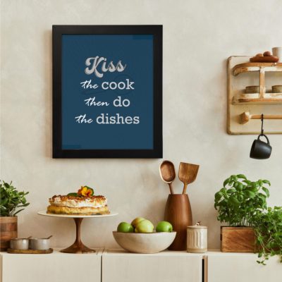 Kiss the cook then do the dishes wall art with white cursive and bold fonts on a dark blue background, perfect for modern kitchens. There is a vegetable table, and above it, there is wall art.