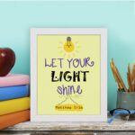 Infidu wall art featuring a lightbulb and the verse Let Your Light Shine from Matthew 5:16 in colorful fonts on a yellow background. displayed on the table in a white frame