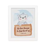 A cute white bear holding a wooden sign with the verse We Love Because He First Loved Us - 1 John 4:19 against a blue sky with clouds. hung on the wall in a white frame