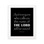 Acts 2:21 Bible wall art featuring bold white text on a black background, reading And everyone who calls on the name of the Lord will be saved. hung on the wall in a white frame