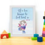 Wall art featuring a joyful bear in winter clothes holding a heart, with the verse We love because He first loved us from 1 John 4:19. displayed on the table in a white frame