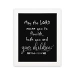 Wall art featuring the quote 'May the Lord cause you to flourish, both you and your children' from Psalm 115:14 on a black background. hung on the wall in a white frame