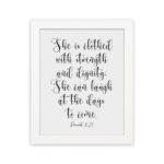 Proverbs 31:25 Christian wall art featuring the text She is clothed with strength and dignity; she can laugh at the days to come. hung on the wall in a white frame