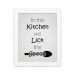 In This Kitchen We Lick the Spoon kitchen wall art featuring black text on a silver-gray background with a minimalist spoon illustration. displayed on the wall in a white frame in a white background