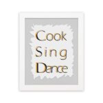 Cook Sing Dance kitchen wall art with black and orange text on a light gray background with jagged white accents. displayed on the wall in a white frame in a white background