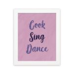 Infidu Cook Sing Dance Kitchen Wall Art with bold navy-blue text on soft pink background, framed in black, modern and lively kitchen design. displayed on the wall in a white frame in a white background