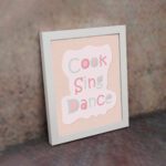 Cook Sing Dance Kitchen Wall Art with pastel pink, green, and blue text on a peach background, perfect for cheerful kitchen decor.