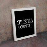 Jesus Christ wall art in white curly font on a black background, showcasing elegant Christian design. displayed on the wall in a white frame