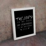 Bible wall art featuring Psalm 18:2 with the text The Lord is my rock, my fortress, and my deliverer in bold and cursive fonts. displayed on the wall in a white frame