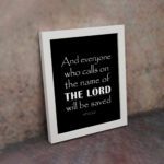 Acts 2:21 Bible wall art featuring bold white text on a black background, reading And everyone who calls on the name of the Lord will be saved. displayed on the wall in a white frame