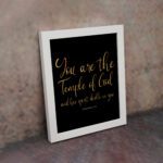 You Are The Temple Of God Bible Wall Art with gold text on a black background from 1 Corinthians 3:16. displayed on the wall in a white frame