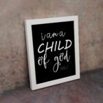 Black wall art with white text reading 'I am a CHILD of god' from John 1:12, showcasing a mix of cursive and bold lettering. displayed on the wall in a white frame