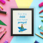 For This Child, We Have Prayed 1 Samuel 1:27 Bible Verse Wall Art with a child on blue bird illustration displayed on the wall in a black frame in a colorful background