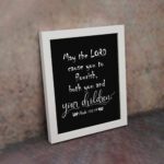 Wall art featuring the quote 'May the Lord cause you to flourish, both you and your children' from Psalm 115:14 on a black background. displayed on the wall in a white frame