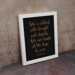 Proverbs 31:25 Christian Wall Art - She Is Clothed With Strength And Dignity, Gold Text on Black Background displayed on the wall in a white frame