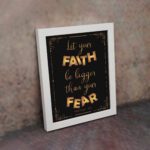 Infidu wall art with the text "Let your faith be bigger than your fear" in gold and bronze, with the Bible verse Hebrews 13:6 on a black background. displayed on the wall in a white frame