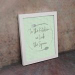 In This Kitchen We Lick The Spoon kitchen wall art with light green background and subtle kitchen utensil pattern in a casual handwritten font. displayed on the wall in a white frame