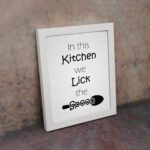 In This Kitchen We Lick the Spoon kitchen wall art featuring black text on a silver-gray background with a minimalist spoon illustration. displayed on the wall in a white frame