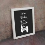 In This Kitchen We Lick The Spoon wall art with white cursive and bold fonts, black background, and stylish cutlery illustrations. displayed on the wall in a white frame