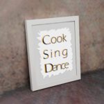 Cook Sing Dance kitchen wall art with black and orange text on a light gray background with jagged white accents. displayed on the wall in a white frame